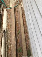 Two railroad rails approximately 6 feet long, laid on dirt and gravel beside a building. Rails are rusty and heavy metal construction.