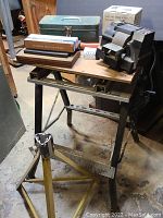 Full view of adjustable metal portable work bench with wooden top, featuring sharpening stones, metal toolbox, and sharpening tools on top. Also shows yellow adjustable metal stand below.