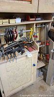 Photo of wood storage cabinet with assorted clamps hanging on/around it and various small boxes of hardware on top.