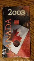 Photo of the original sealed packaging showing the '2003 CANADA' text and maple leaf design with the coin visible through the case.