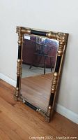 Full view of rectangular antique mirror leaning against white wall, showing black and gold wooden frame with carved corners and sections.