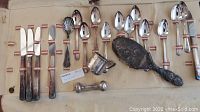 Photo showing the collection of antique sterling silver and silver plate items including baby rattle, cup, brush handle, knives, spoons and hand mirror.