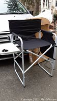 Black canvas director's chair with aluminum frame, snack tray with cup holder, and side pocket storage shown outdoors beside a vehicle.