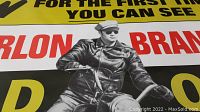 Close-up photo of the center of the poster showing Marlon Brando riding a motorcycle in black leather jacket and cap, bold red text