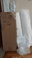 Box labeled '10x10 Tent' with white wrapped metal poles and folded canopy fabric alongside it on the floor.