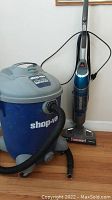 Blue and gray Shop-Vac 14-gallon wet/dry vacuum and blue Bissell vacuum/steam mop side by side against a wall on hardwood floor.