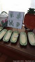 Photo of six ceramic corn holders in green and yellow, displayed on a wooden surface with a white ceramic cookie jar with pink tulip design, a square ceramic plate with herb illustrations, and a ceramic pineapple-shaped container.