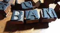 Close-up of wooden block stamps showing letters B, A, M with black rubber surfaces worn with cracks and some vinyl loss.