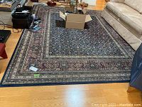 Full view of the larger rug placed on wood flooring next to sofa showing overall pattern and condition with visible wear and soiling