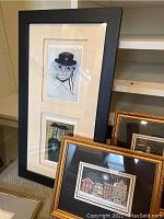 Framed prints under glass with a tall black frame containing two prints of a cat or sheep wearing a hat and glasses, gold framed architectural scenes visible.