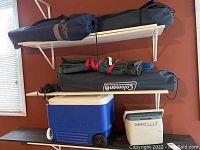 Shelf showing 4 camp chairs in bags stacked above 2 coolers, one Igloo and one Coleman Personal 8.