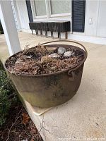 Cast iron kettle showing rust, dirt, rocks, and dried plant material inside. Positioned on concrete porch.