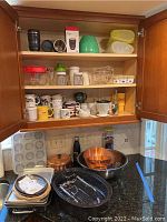 Full view of kitchenware inside cabinet and on countertop showing mugs, glassware, roasting pans, copper bowls, enamel roasting pan and cast iron plates.