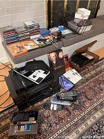 Overview of lot items on carpet and bench including VHS players, remotes, CDs, DVDs, cassette tapes, booklets, iPod, and cables.