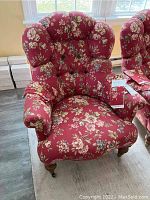Front angled view of the red floral upholstered armchair showing tufted back, armrests with fabric covers, and wooden front legs on casters.