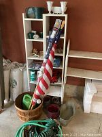 Photo of white wooden shelves holding two white milk glass-style outdoor candle holders, glass jars, small household items, and plastic pots. To the left area is a large woven wooden basket containing American flags, a green watering container, and several small pots. Garden hoses and tools lay on the floor in front.