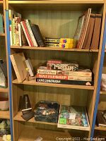 Wooden shelving unit with multiple shelves containing books and board games, a metronome, and rolls of book covers.