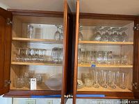 Wooden cabinet open showing multiple shelves filled with assorted glassware including stemware, drinking glasses, plates, and bowls.