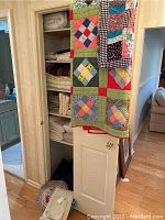 Wide view showing hand stitched quilt with green borders and colored patchwork, open closet with neatly folded bedding and blankets, door partially open with hanging quilts.