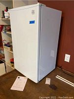 Front and side view of the closed white mini fridge, showing a blue sticker on the door and some minor marks on the side.