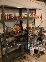 Two metal shelving units housing assorted copper and metal kitchenware including kettles, pots, trays and bowls spread across multiple shelves.