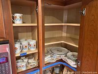 Six dinner plates stacked with ten lunch plates, mugs, coasters, napkins, and tea bag plate arranged on shelves in wooden cabinet.