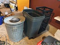 Metal and plastic trash cans with lids and three black plastic milk crates in a cluttered room environment