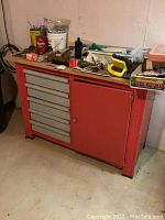 Red metal framed workbench with five beige plastic drawers on left, cabinet door on right, and pressed board top with assorted items on it in a basement setting.