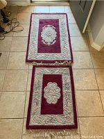 Two burgundy and beige floral area rugs with fringed edges on tiled floor.