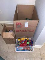 Clear plastic box with lid filled with crayons, colored pencils, and art supplies, positioned in front of the cardboard boxes on the floor.