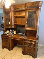 Full frontal view of the large wooden desk with hutch. Two glass doors with decorative framing on the upper portion. Open shelving in the middle. Desktop cluttered with items. Curved drawer fronts in the lower section.