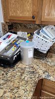 Photo showing various white plastic baskets and black plastic baskets on kitchen countertop with a tall transparent jar with blue lid and kitchen utensils in a plastic holder.