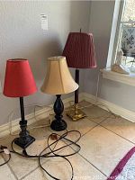 Three table lamps with various base materials and shades placed on tile floor near wall and window.