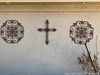 View of three metal wall hangings mounted on stucco exterior wall: two circular floral patterned pieces and one cross shape in the center.