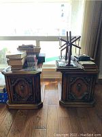 Pair of dark wooden side tables with carved doors shown with books and other items on top, set against window light.