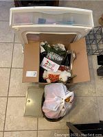 White plastic underbed storage drawer with transparent front panel, cardboard box with winter decorations, small metal lockbox, and a trash bag in front.