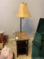 Full view of the wooden side table with the marble top and the lamp with ivory shade turned off, placed beside a green upholstered chair.