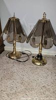 Two vintage table lamps with curved smoky glass panel shades and brass bases placed on carpeted floor, showing lamp structure and visible glass damage.