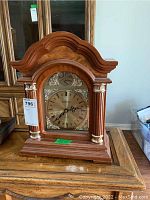 Front view of wooden mantel clock with arched top and decorative columns, clock face with Roman numerals and silver ornamental accents