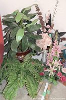 Wide view of lot showing large artificial green plant with broad leaves, several artificial flower stems and arrangement, and fern-like artificial plant in front.