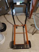 Front view of metal dolly showing black handle, orange metal frame, and large wheels.