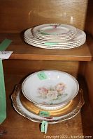 Stack of dinner and dessert plates on upper shelf, showing delicate rose floral design with gold trim.