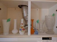 Photo shows a group of vases on a shelf including white milk glass vases with hobnail and floral design, one orange ribbed vase, and a clear cut glass vase partially visible.