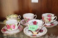 Photo of five floral tea cups and matching saucers in display cabinet, showing variety of pink and red rose patterns with gold trim