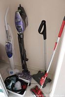 Three upright cleaning devices: a white and blue H2O steam cleaner with accessories in a bin on the floor, a tall purple and black Shark carpet and bare floor cleaner, and a black Shark battery-powered swivel sweeper base leaning against the wall.
