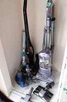Group photo showing Shark Navigator vacuum with hose, Kenmore stick vacuum, Omega mini steam zapper, and assorted vacuum attachments on floor in corner.