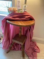 Wood round accent table with pedestal base covered with pink fabric, showing entire table and base.