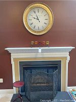 Gold-toned wall clock above fireplace with black metal fireplace screen and small red candlestick end table below