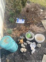 Wide view of assortment of garden items on ground by wall including painted metal milk jug, flowerpots, ornaments, and banner holders.