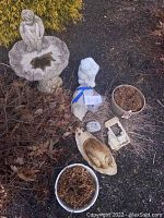 Full lot view of garden ornaments including birdbath, female figure, cat, snail, pots, decorative rock and tile in garden bed.